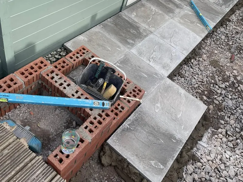 Brick step being built beside newly laid grey paving slabs, with a spirit level and tools