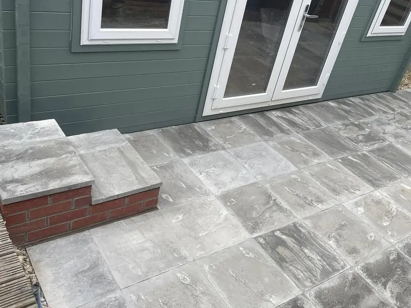 Finished grey slab patio with a brick-built step up to the doors of a green garden room