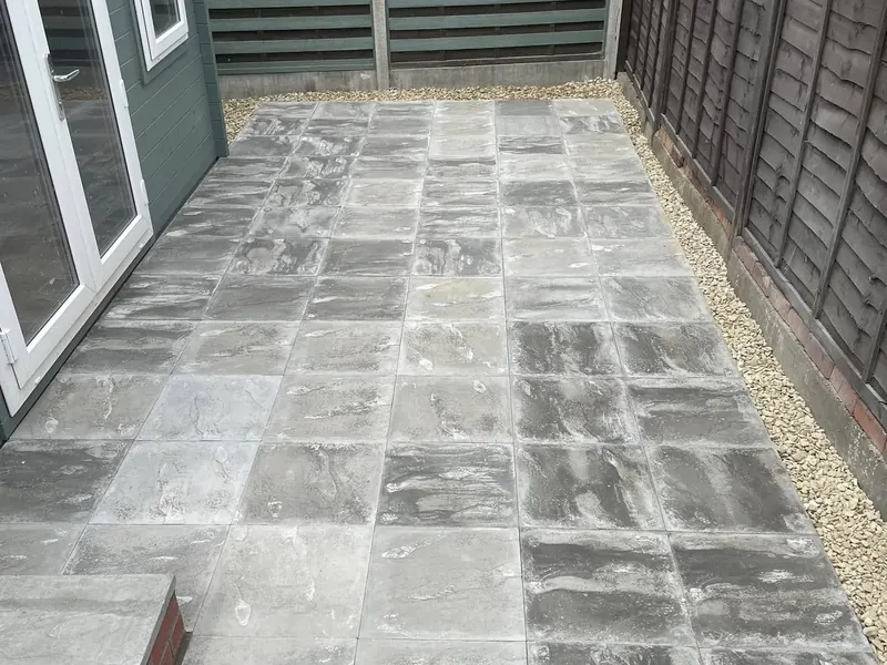Grey paving slabs laid as a patio with a gravel border, between a green garden room and a fence